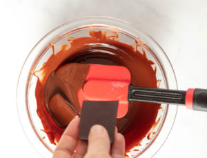 someone adding a piece of chocolate to a glass bowl with melted chocolate