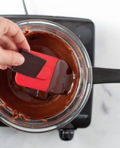 Someone adding a piece of Chocolate bars to a bowl with melted chocolate over a pot of water on the stove